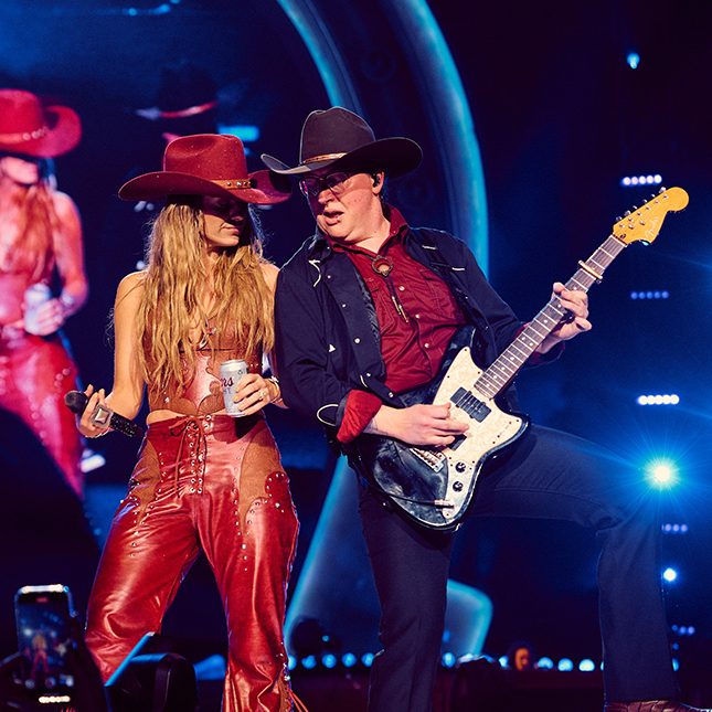 Female singer in a cowboy hat on a lit up stage with a guitarist.