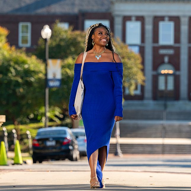 Student in blue dress walks down College Ave at UNCG with the Curry building behind her.