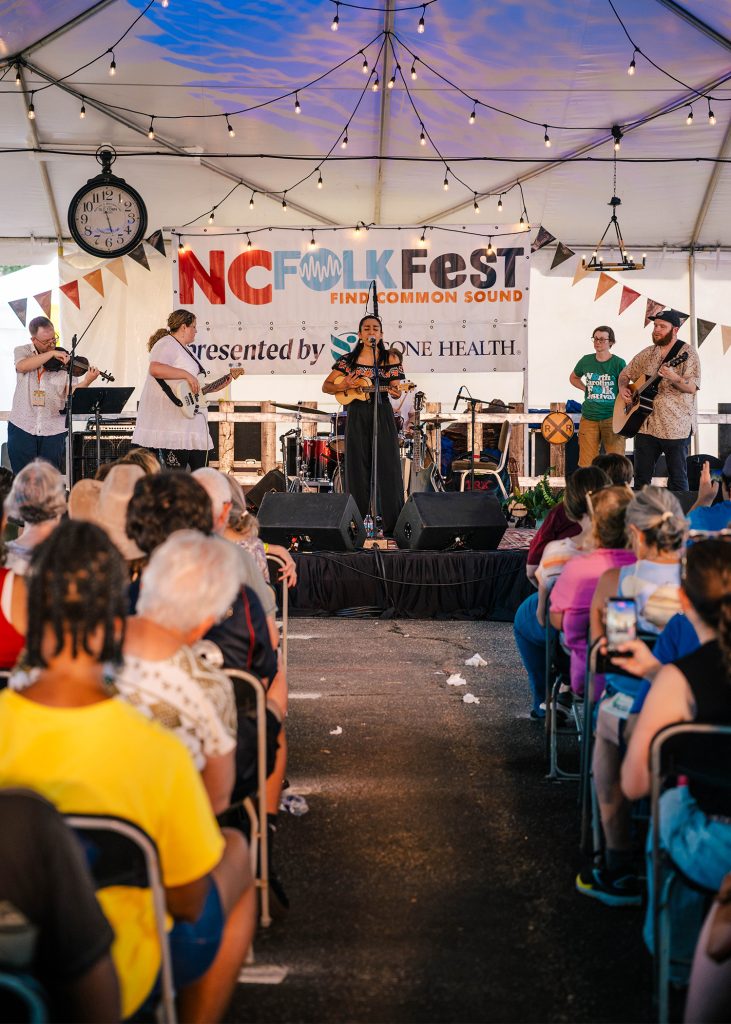 Band plays under a Folk Fest tent with a packed audience in the foreground.