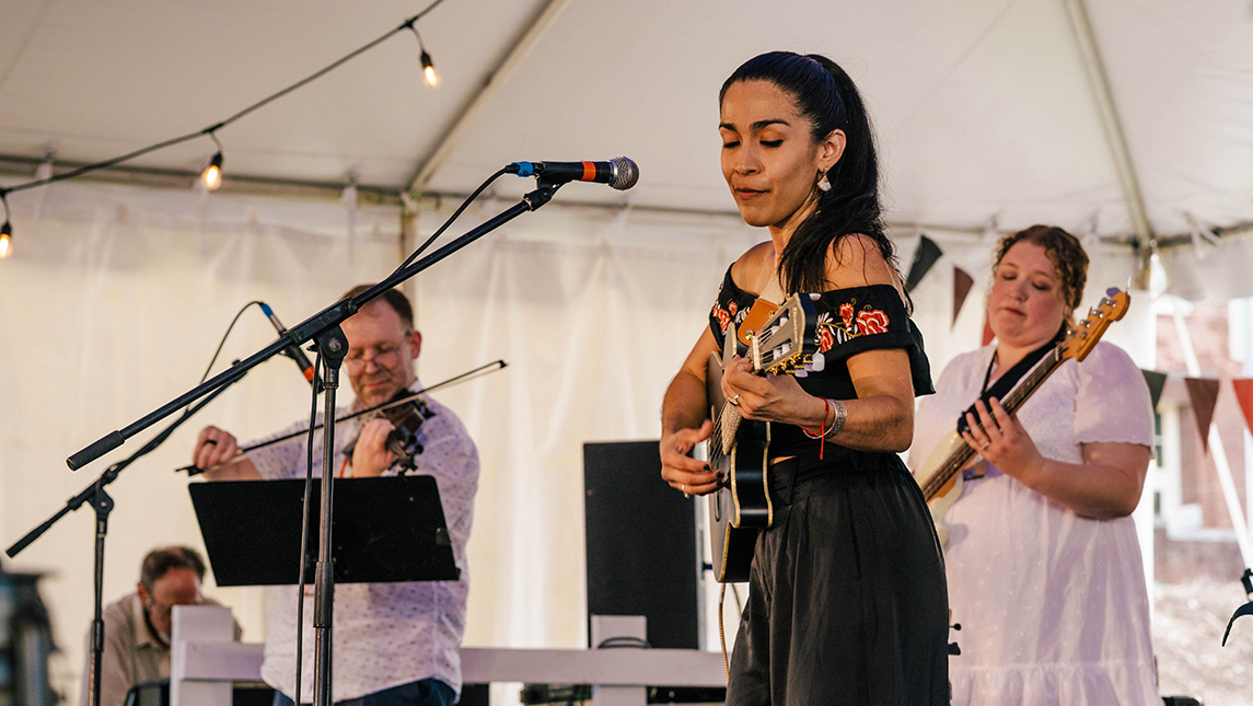 Woman plays guitar on a stage under a tent with a violinist and bass player behind her.