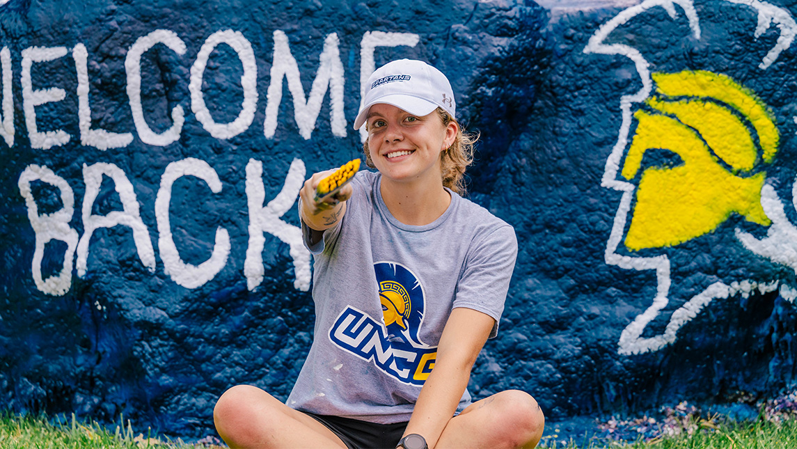 Student sits in front of a painted rock with Welcome Back & UNCG logo on it. She's pointing to the camera with her paintbrush.
