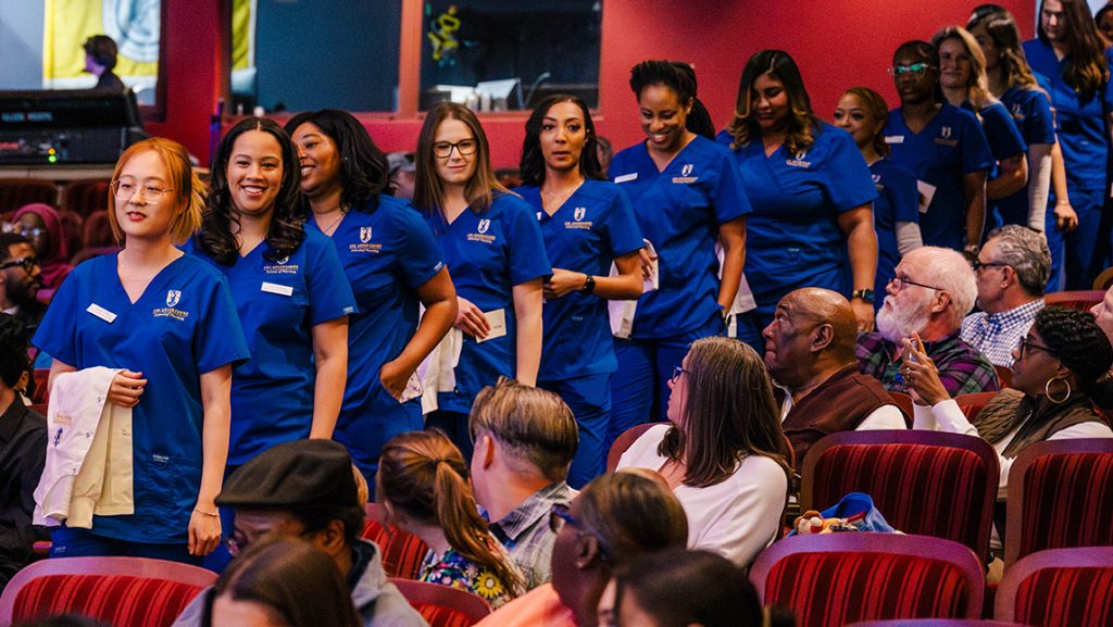 UNCG nurses in scrubs with white coats in their arms, file into auditorium as family members look on.