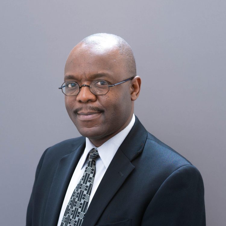 Professional studio headshot of Morgan Chitiyo wearing a dark suit, patterned tie, and glasses against a plain gray background.