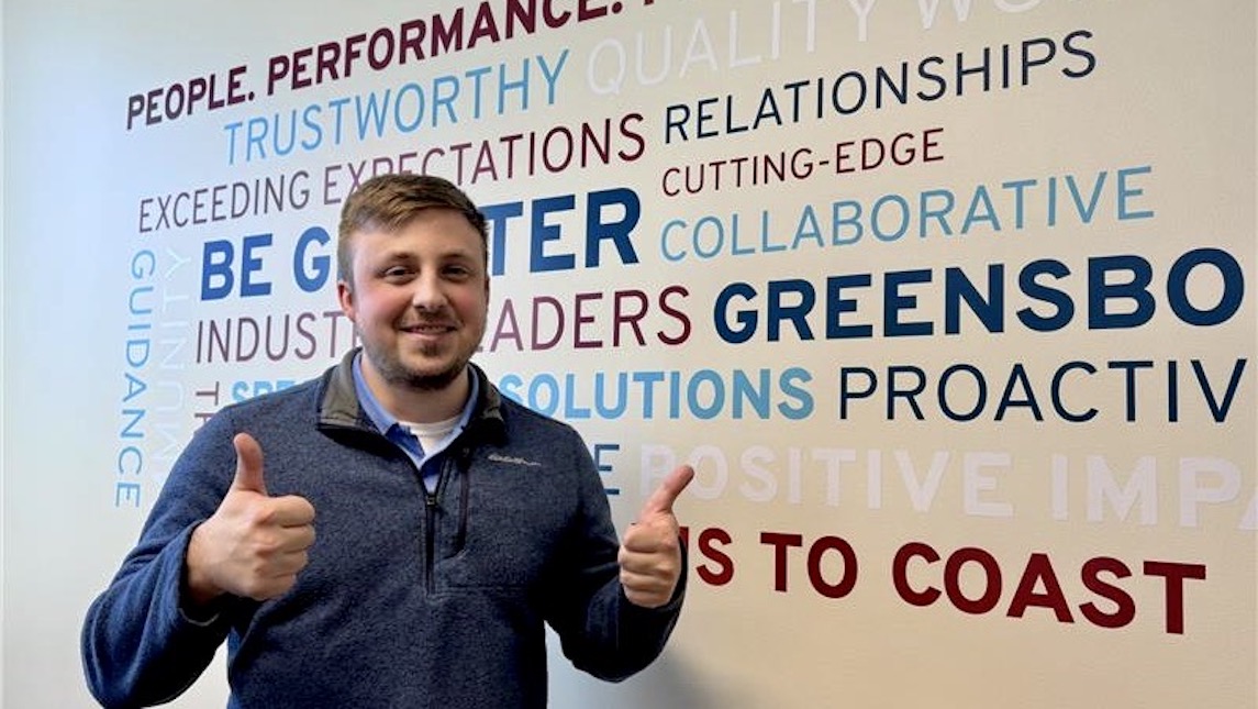 UNCG BSBE alumnus Noah Brabble standing in front of a motivational board.