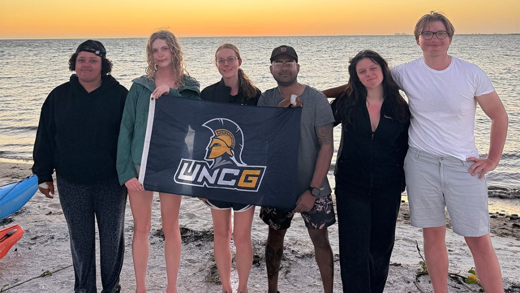 Students hold a UNCG flag on a beach.