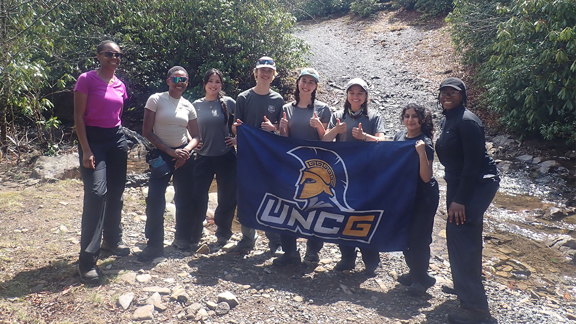 Hikers hold a UNCG flag next to a stream.