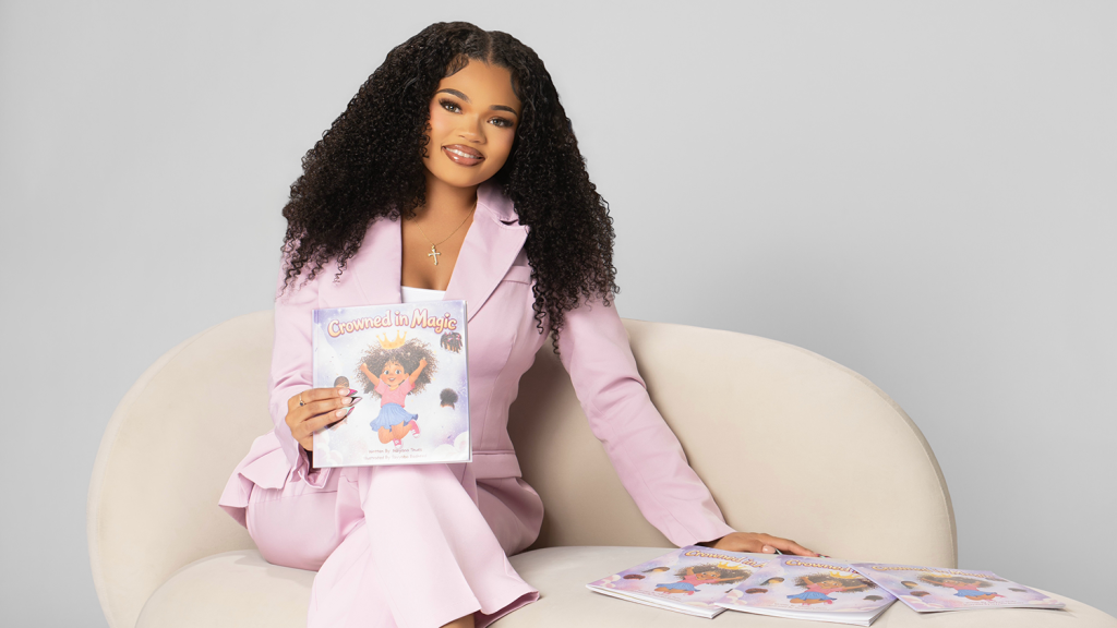 Naiyana Thues sits on a cream-colored chair holding Crowned in Magic, with more copies beside her.