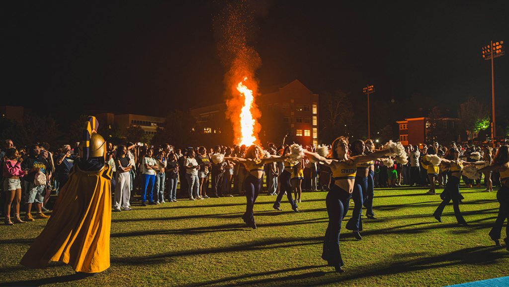 Bonfire on campus recreation field with a crowd dancing and cheerleaders and Spiro in the foreground.