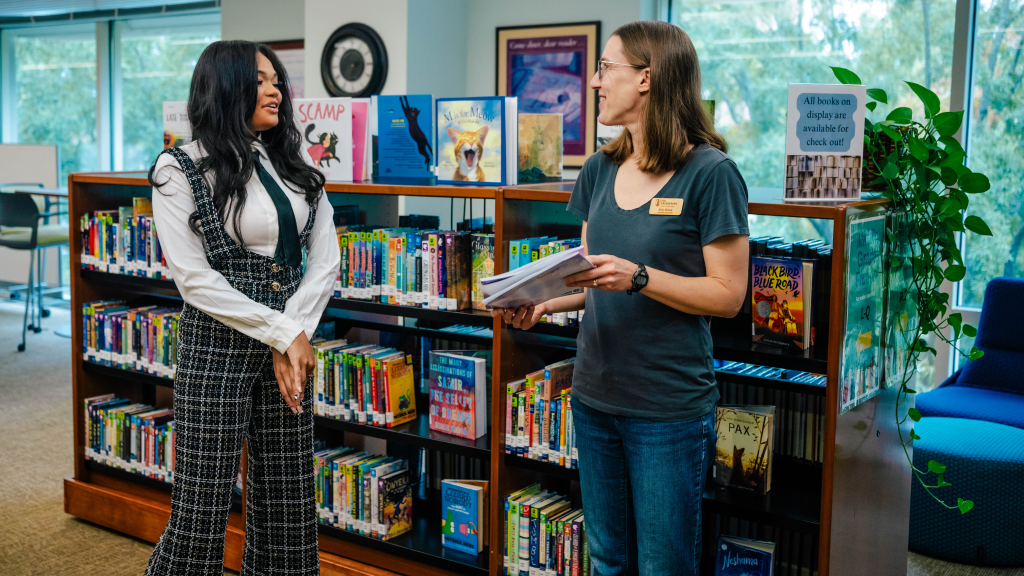 Author Naiyana Thues hands copies of Crowned in Magic to a librarian among the children’s bookshelves.