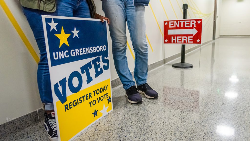 An election sign rests on the floor at people's feet.