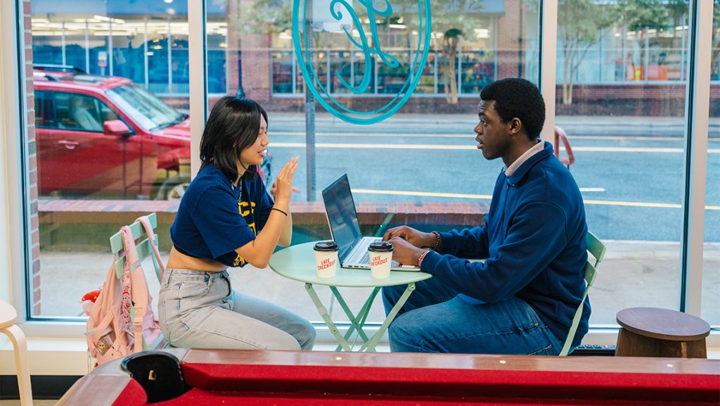 Two people sit at a table with their backpacks and laptops and coffee cups.
