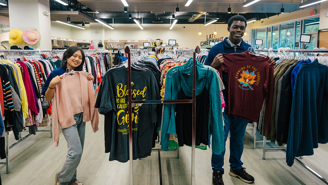 Two UNCG students hold up shirts on hangers in the middle of a store.